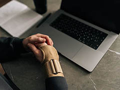 Close-up top view of male hands wearing wrist brace, massaging painful wrist while working remotely from home office, surrounded by laptop and notebook.